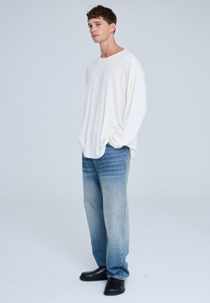 Camicia bianca a maniche lunghe, vestibilità morbida, tessuto morbido; jeans dritti azzurri chiari, leggermente sbiaditi; scarpe nere slip-on, design semplice.