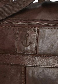 Sac en cuir marron à finition texturée, doté d'une fermeture éclair et d'un patch de marque avec un motif d'ancre. Accents cousus visibles.