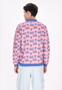 Giacca rosa con zip e colletto blu, caratterizzata da un motivo con palme e isole. Texture liscia con polsini e vita a coste blu.