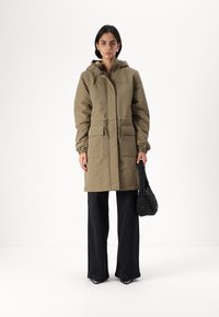 Khaki parka s kapucí, vybavený dvěma předními kapsami, elastickými manžetami a stahovacími šňůrkami v pase. V kombinaci s černými kalhotami širokého střihu a pletenou taškou.