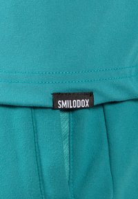 Tela verde azulado con textura suave, que presenta un borde doblado y una etiqueta negra con texto blanco que dice "SMILODOX" en la parte inferior.