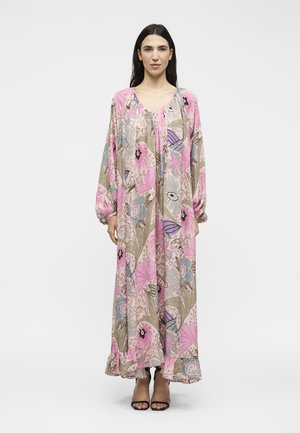 Γυναίκα με μακριά σκούρα μαλλιά, φορώντας ένα μακρύ, άνετο φόρεμα με λουλουδάτο σχέδιο σε ροζ, μπεζ και μπλε αποχρώσεις, συνδυασμένο με μαύρες ανοιχτές γόβες, στέκεται σε λευκό φόντο.