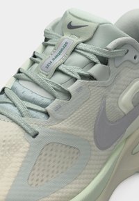 Κοντινό πλάνο ενός ανοιχτό πράσινου αθλητικού παπουτσιού Nike με υφασμάτινη υφή, κορδόνια και εμφανής ετικέτα "Structure Plus" στη γλώσσα.