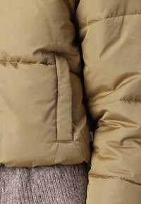 Vaaleanruskea puffatakkia, jossa on sileä pinta ja pystysuuntainen tikkauskuvio, sivutasku sekä vaalea ruskea neulepusero sen alla.