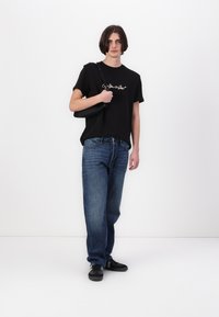 T-shirt en coton noir avec logo script blanc, jeans en denim bleu et baskets noires. Le modèle porte un sac bandoulière noir. Fond uni.