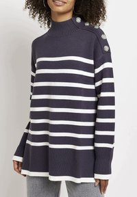 Femme portant un pull à rayures horizontales bleu marine et blanches avec des boutons argentés sur l'épaule et des poignets côtelés.