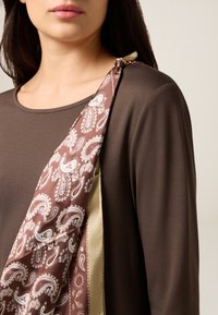 Bruine top met lange mouwen en een zijden sjaal met paisley-patroon in bruin en wit, los over een schouder gedrapeerd en vastgemaakt met een beige lint.