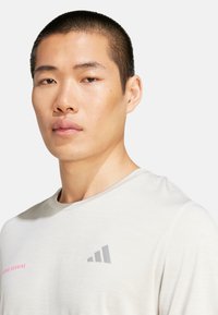 T-shirt de sport gris clair à manches courtes, avec un texte rose "adidas running" et un logo gris sur le côté gauche. Texture lisse.