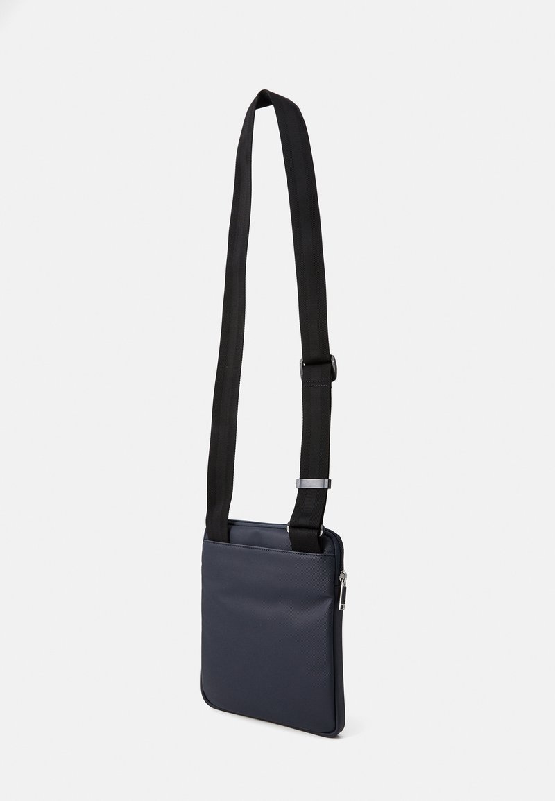 Bolso cruzado azul marino hecho de material suave, con una correa negra ajustable, cierre de cremallera y un diseño rectangular minimalista.