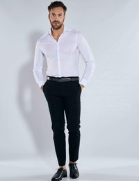 Weißes Langarmhemd mit Kragen, kombiniert mit schwarzen, eng anliegenden Hosen und schwarzen Schuhen, präsentiert vor einem hellgrauen Hintergrund.