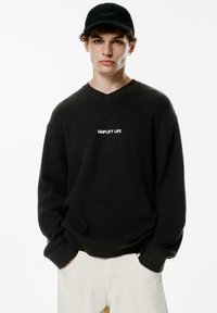 Pulover negru tricotat cu decolteu în V și mâneci lungi. Caracterizat prin textul alb "SIMPLIFY LIFE" pe piept. Asortați cu pantaloni de culoare deschisă.
