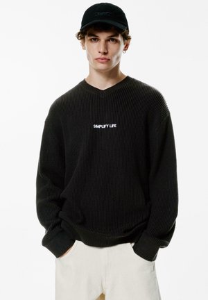 Sort strikket sweater med V-hals og lange ærmer. Har hvid "SIMPLIFY LIFE" tekst på brystet. Kombiner med lyse farvede bukser.