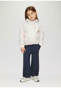 Leichte, flauschige Zip-Jacke in Pastellfarben; marineblaue Cargo-Hosen, weiße und graue Sneaker mit Mesh-Akzenten; Mädchen steht mit Händen in den Taschen.