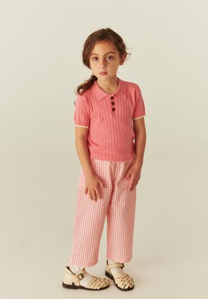Young girl standing wearing a pink short-sleeve knit top, pink houndstooth wide pants, white socks, and beige woven sandals.