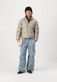 Puffer jacket in light grey, featuring a high collar and zip closure. Paired with loose-fitting blue denim cargo pants and black shoes.