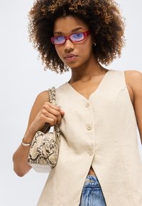 Femme aux cheveux bouclés portant des lunettes rouges et un haut sans manches beige boutonné, tenant un petit sac à main en cuir de serpent, sur un fond uni.