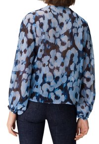 Femme portant un chemisier transparent à manches longues avec un motif floral abstrait bleu et noir, associé à un jean bleu foncé, vue de dos.