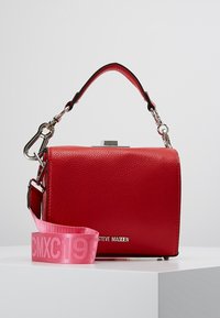 Sac à main en similicuir rouge avec une forme structurée, des ferrures argentées et une sangle rose amovible portant le texte "CMXC19".