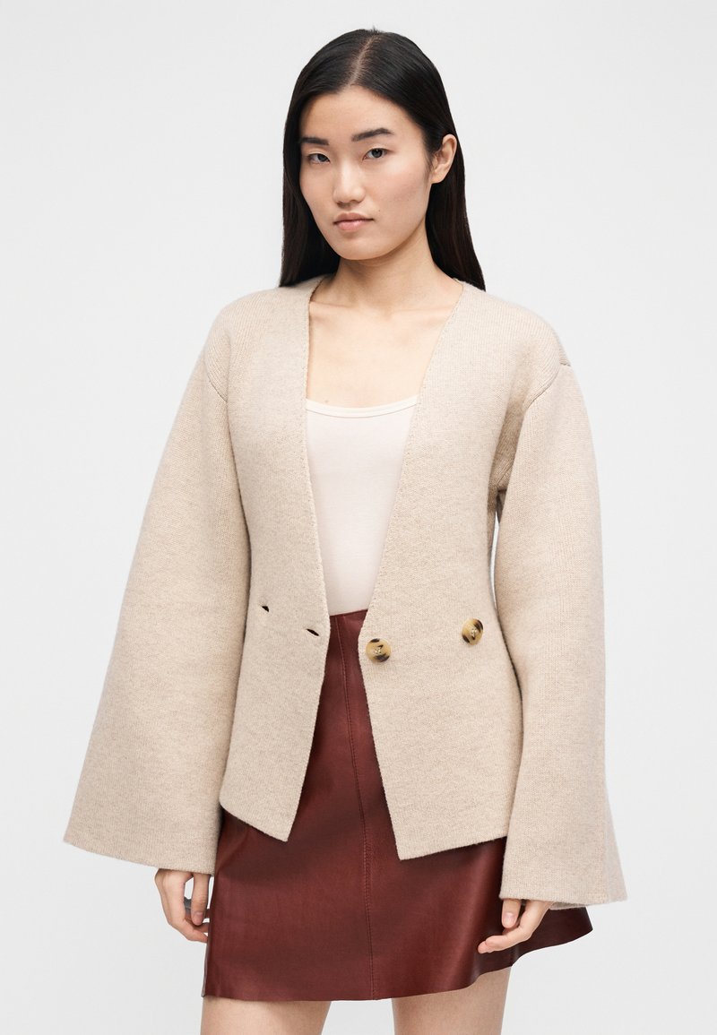 Beige cardigan i uldblanding med brede ærmer og V-udskæring, med to store knapper og minimalistisk design. Kombineret med en brun lædernederdel.