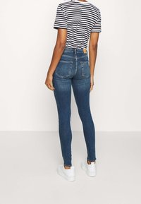 Jeans ajustados de mezclilla azul oscuro, de cintura alta, con dos bolsillos traseros, combinados con una camiseta de rayas en blanco y negro y zapatillas blancas.