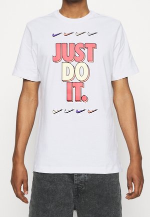 T-shirt en coton blanc avec un texte audacieux "JUST DO IT" en rose et jaune, accompagné de plusieurs logos Nike en dessous dans différentes couleurs. Manches courtes, col rond.