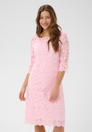Mlada žena s dugom smeđom kosom, odjevena u ružičastu čipkastu haljinu do koljena s rukavima tri četvrtine, smiješi se na jednostavnoj pozadini.