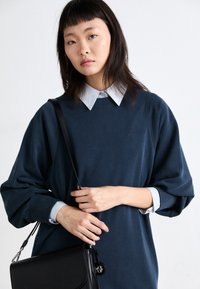 Μπλε ναυτικού oversized φούτερ από μαλακό ύφασμα, με στρογγυλή λαιμόκοψη, μανίκια πουκάμισου σε ανοιχτό μπλε με μανσέτες και μια μαύρη τσάντα χειρός με κυκλικό διακοσμητικό.