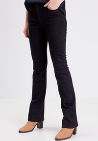 Jeans negros acampanados con una textura suave, que presentan bolsillos frontales y costuras mínimas. Combinados con botas de tacón marrón claro.