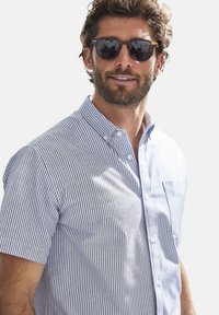 Kurzarmhemd mit Knopfverschluss in Blau- und Weißstreifen, mit einer Brusttasche und einem klassischen Kragen. Getragen mit einer dunklen Sonnenbrille.