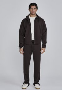 Bruine zip-up jas met capuchon, gecombineerd met bijpassende broeken. Stof lijkt zacht en textuur te hebben. Witte t-shirt eronder, sneakers aan de voeten.