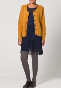 Gilet en tricot jaune moutarde avec un col rond et une fermeture à bouton, superposé sur une robe bleu marine, associé à des collants gris et des chaussures noires.