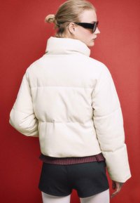 Weiß gesteppte Pufferjacke mit hohem Kragen, geripptem bordeauxfarbenem Saum, kombiniert mit schwarzen Shorts vor rotem Hintergrund.