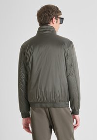 Giacca imbottita verde oliva con colletto alto, polsini elasticizzati e orlo. Finitura liscia con cuciture visibili, indossata sopra pantaloni abbinati.
