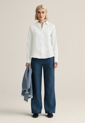 Lyshåret kvinde, stående, iført hvid skjorte med knapper, brede blå bukser, holder blåt frynset tørklæde i venstre hånd mod neutral baggrund.