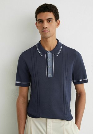 Mladý muž ve tmavě modrém polokošili s krátkým rukávem a bílým lemováním, stojící před jednobarevnou světlou zdí, ruce volně podél těla.