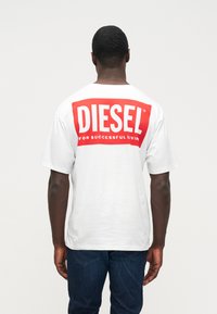 Άνδρας που φοράει λευκό T-shirt της Diesel με μεγάλο κόκκινο λογότυπο και σλόγκαν "Για Επιτυχημένη Ζωή", γυρισμένος προς τα πίσω, συνδυασμένο με μπλε τζιν σε απλή φόντο.