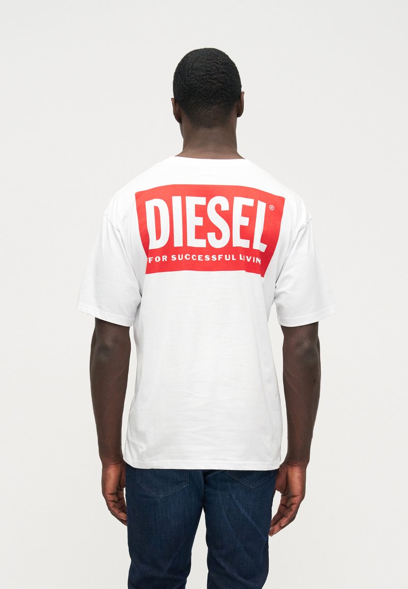 Άνδρας που φοράει λευκό T-shirt της Diesel με μεγάλο κόκκινο λογότυπο και σλόγκαν "Για Επιτυχημένη Ζωή", γυρισμένος προς τα πίσω, συνδυασμένο με μπλε τζιν σε απλή φόντο.