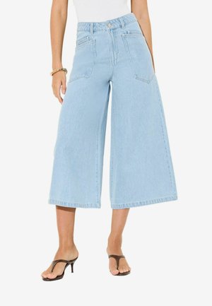 Vrouw draagt lichtblauwe denim jeans met wijde pijpen en cropped stijl, witte top, bruine sandalen met hak en een gouden armband om haar linkerpols.