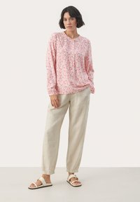 Långärmad rosa blus med ett lövmönster, rund halsringning och knapppdetalj, tillsammans med lösa beige linnebyxor och vita sandaler.