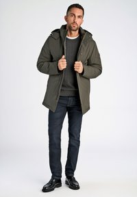 Mænds olivengrønne polstret jakke med hætte, frontlynlås og trykknapper, båret over en mørkegrå kabelstrikket sweater og mørke jeans.