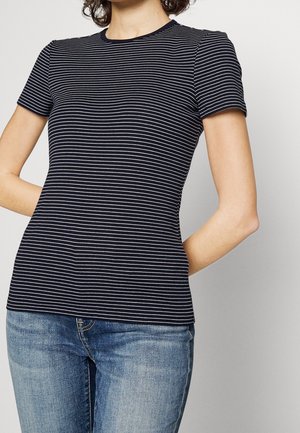 Woman wearing a fitted black t-shirt with thin white horizontal stripes and blue jeans, standing with hands behind her back.