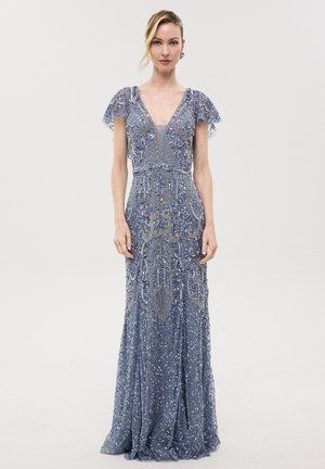 Femme portant une robe longue bleu-gris avec un travail de perles fin, des manches courtes en dentelle et un décolleté en V profond, debout devant un fond uni.