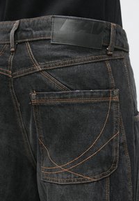 Črne kavbojke iz jeansa z oranžnim šivanjem, pravokotno usnjeno etiketo na pasu in stranskim žepom z dekorativnim šivanjem.