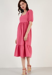 Puff-sleeve, neliönmuotoinen vaaleanpunainen mekko, jossa on kerrostettu helma. Silkkinen kangas, polvimitan leikkaus, yhdistettynä monivärisiin remmikenkiin.