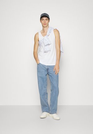 Weißes Tanktop, blaue Jeans und weiße Sneaker. Ein gestreiftes blau-weißes Hemd hängt über der Schulter, dazu eine blaue Mütze.
