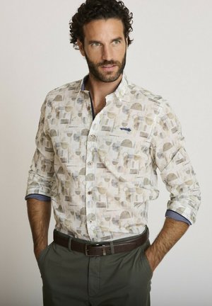 Homme aux cheveux bouclés foncés et à la barbe, portant une chemise claire à motifs boutonnée, un pantalon vert et une ceinture marron, les mains dans les poches.