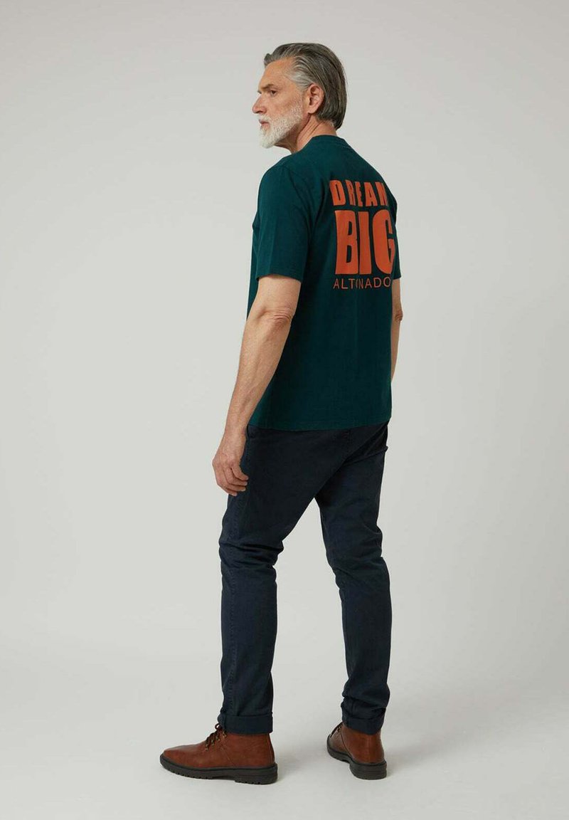 Camiseta de algodón verde oscuro con "DREAM BIG" en naranja en la parte trasera. El modelo lleva pantalones azul marino y botas marrones.