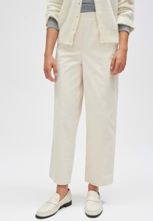 Person trägt cremefarbene Hosen mit weitem Bein, weiße Loafers, graues Langarmshirt und einen cremefarbenen, knöpfbaren Cardigan vor einem einfarbigen Hintergrund.