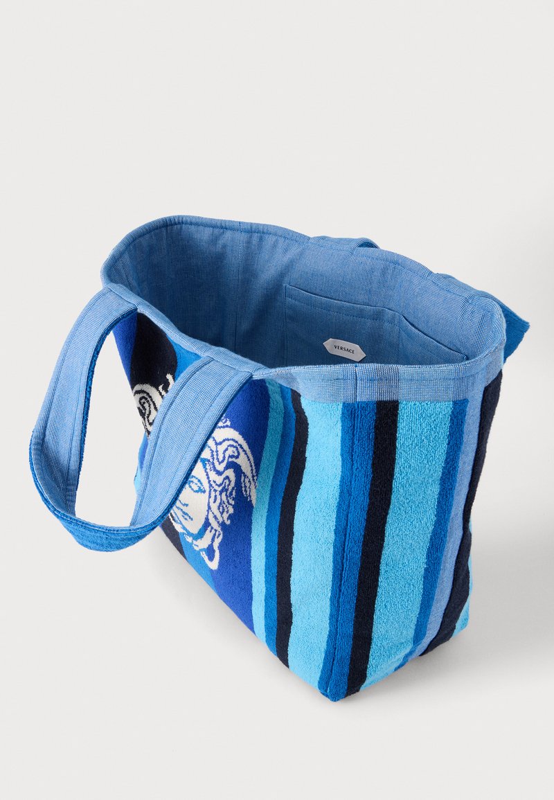 Sac cabas rayé bleu avec de larges anses et un motif latéral représentant une tête de Méduse, avec une poche intérieure portant l’étiquette Versace.
