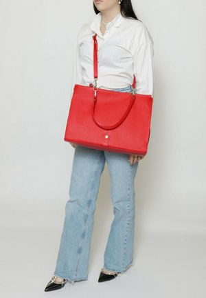 Persona che indossa una camicia bianca e jeans azzurri tiene una grande borsa rossa con tracolla e manici.
